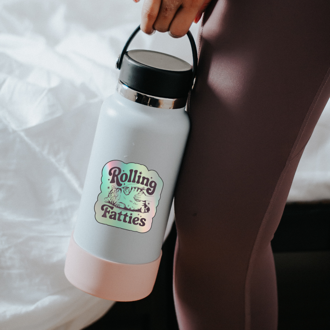 Holographic sticker with a hand rubbing a fat, relaxed cat and the phrase “Rolling Fatties” in playful black text