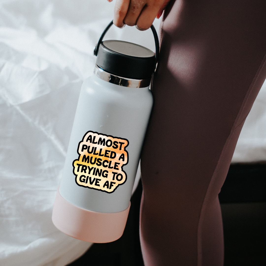 Holographic sticker that reads “Almost pulled a muscle trying to give AF” in bold black text over a peach-pink sparkle background