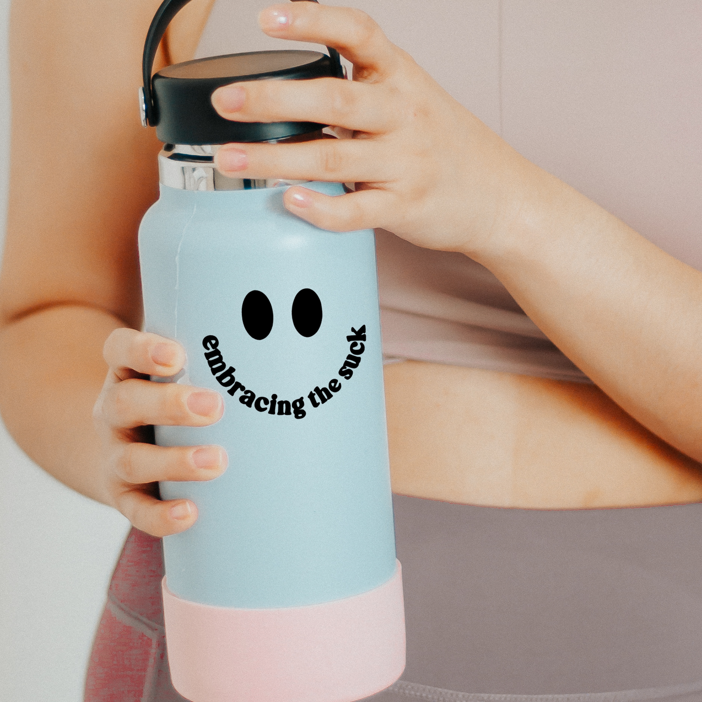 Hands holding a blue water bottle with a smiley face decal that says embracing the suck