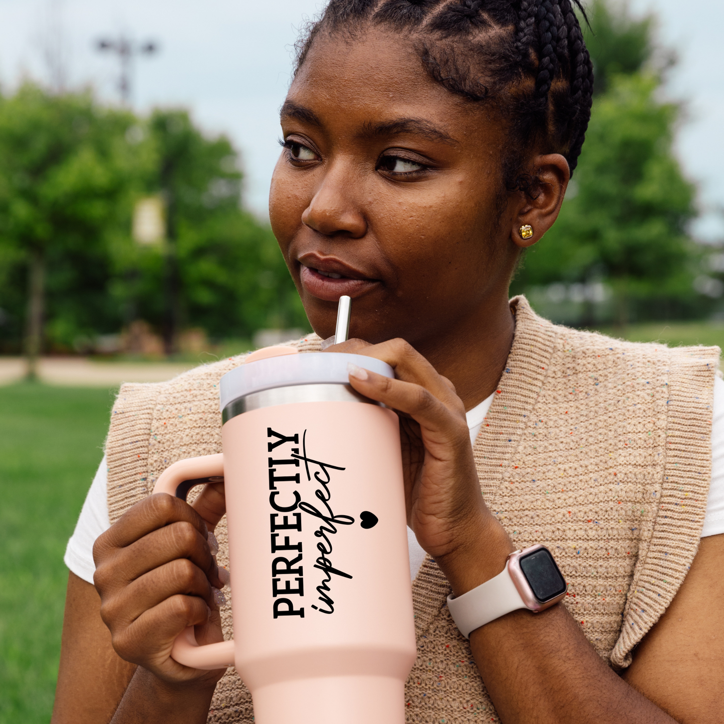 Woman drinking from a pink insulated mug with 'Perfectly Imperfect' text in a park.