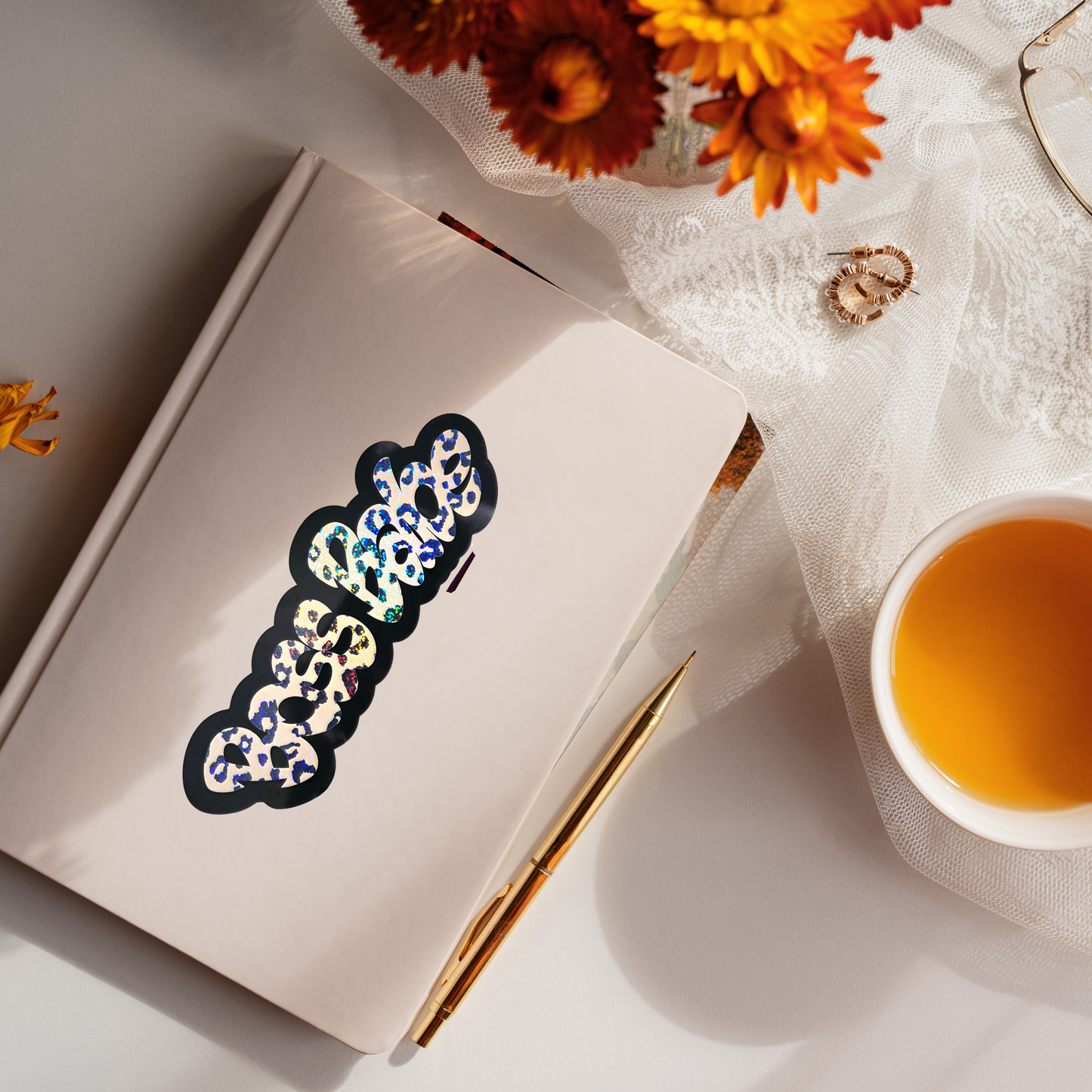Notebook with decorative Boss Babe sticker, a cup of tea, and flowers on a light surface
