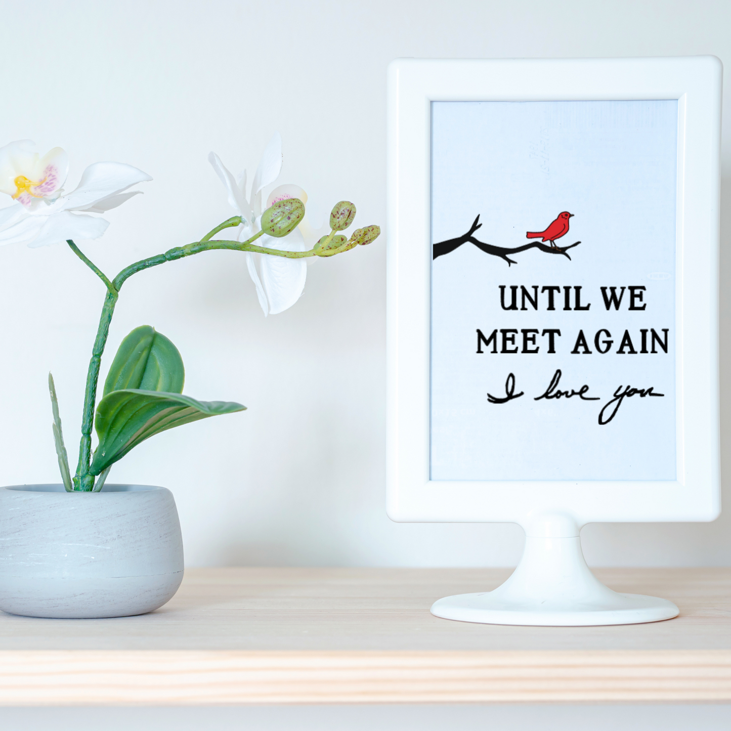 White-framed print on shelf beside orchid plant, featuring a red cardinal on a branch and the text “Until we meet again. I love you” in black script.