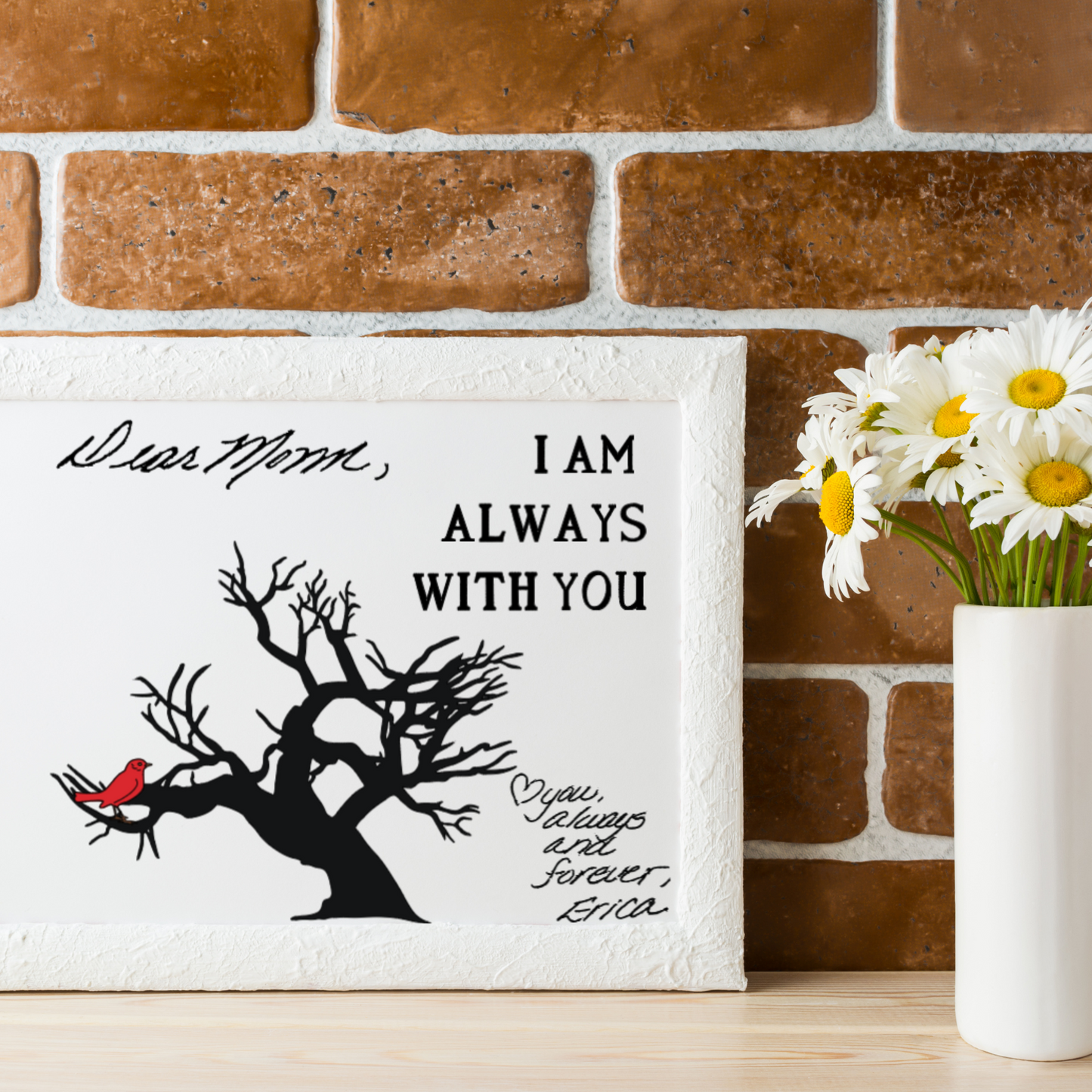 Framed memorial artwork with a black tree and red cardinal, featuring handwritten text that says “Dear Mom,” and “I am always with you. Love you, always and forever, Erica,” displayed on a wood shelf with a white vase of daisies against a brick wall.