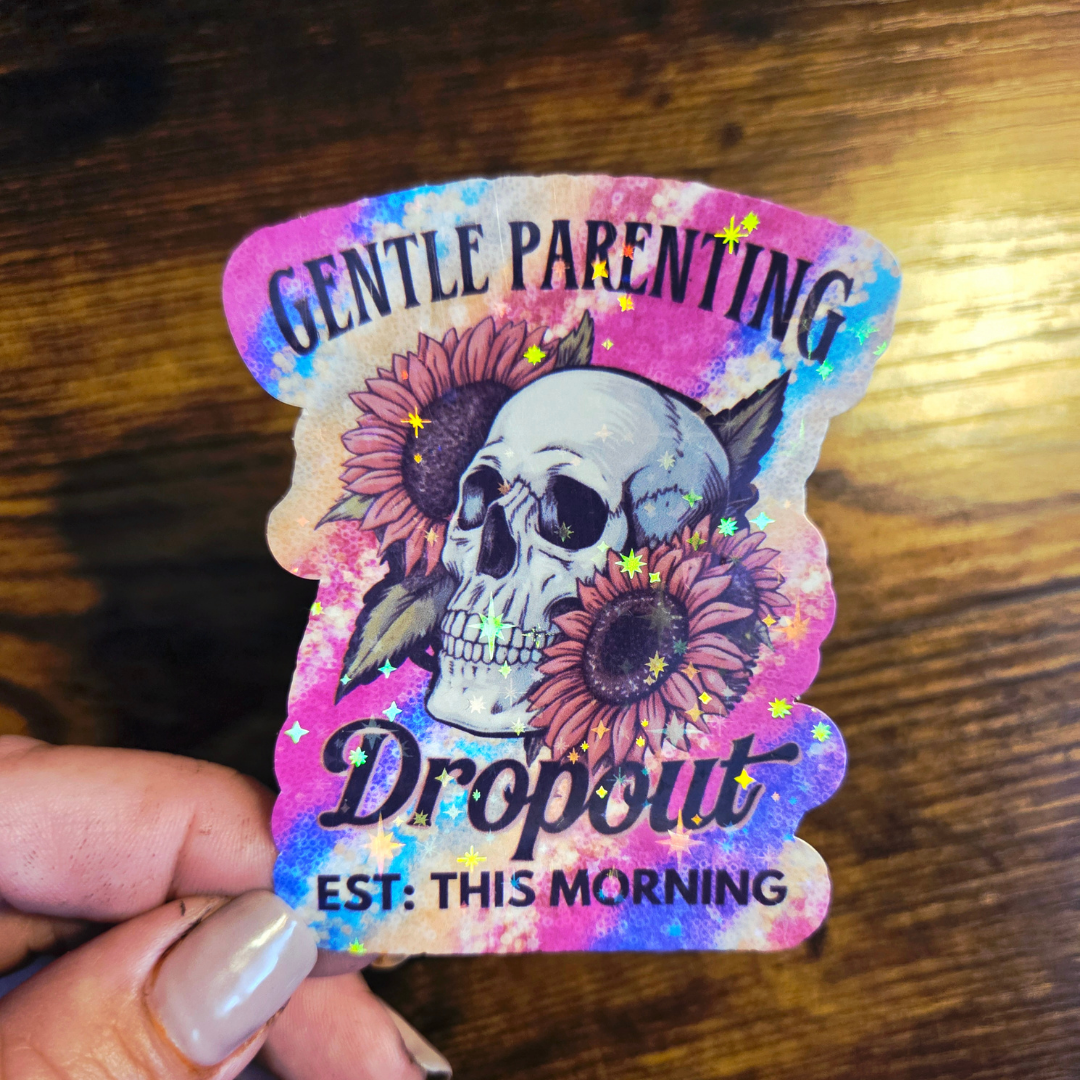 Sticker with skull design and text 'Gentle Parenting Dropout EST: This Morning' held by a hand on a wooden surface.