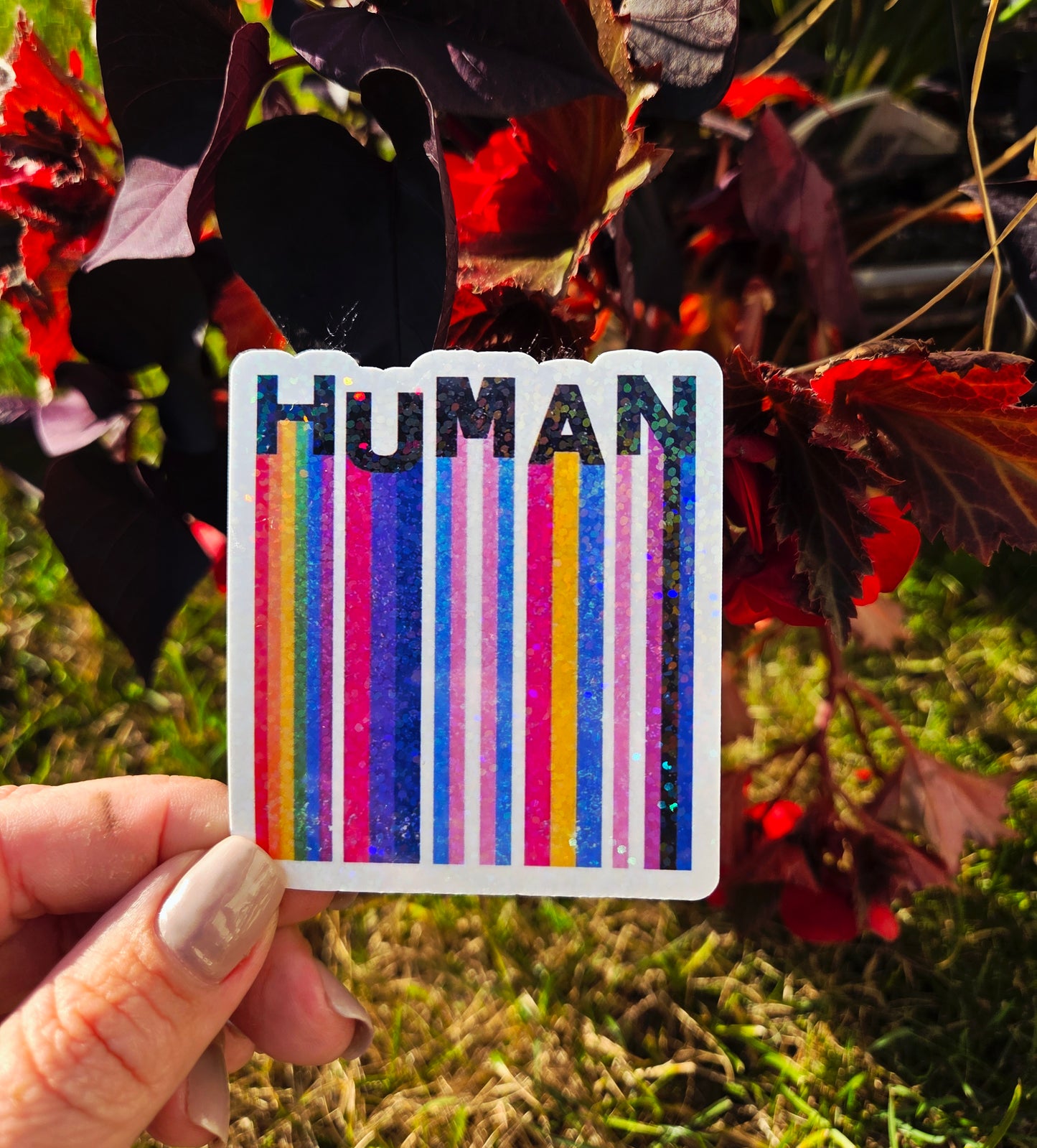 Square holographic sticker with rainbow barcode stripes and the word “HUMAN” in bold black text