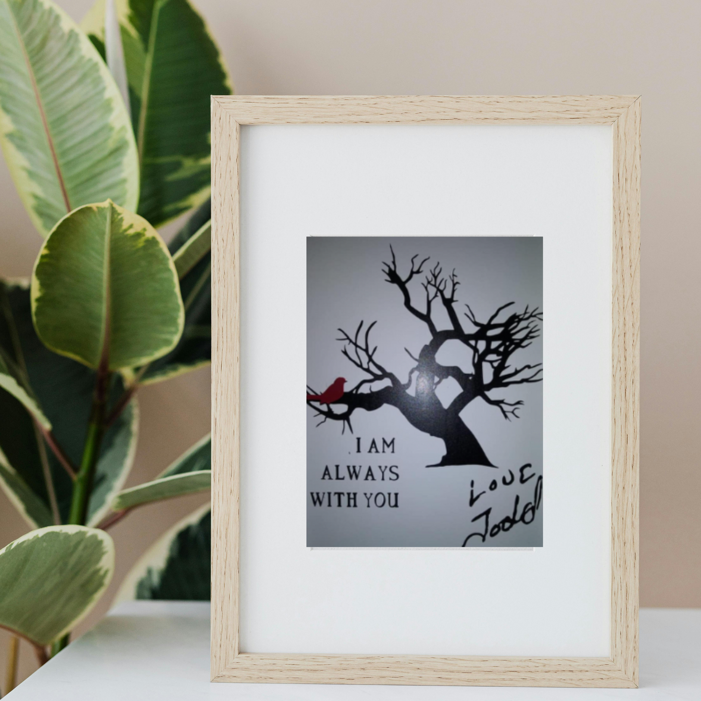 Framed memorial print featuring a black tree and red cardinal with the message “I am always with you” and a handwritten “Love, Todd” signature, placed on a white shelf beside a large leafy plant.