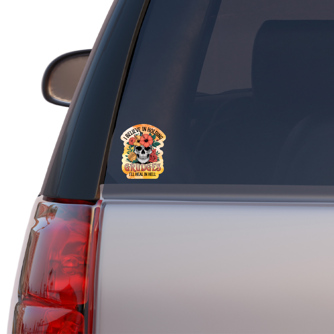 Holographic sticker with a floral-decorated skull and bright orange accents. Text reads “I Believe in Holding Grudges – I’ll Heal in Hell.”