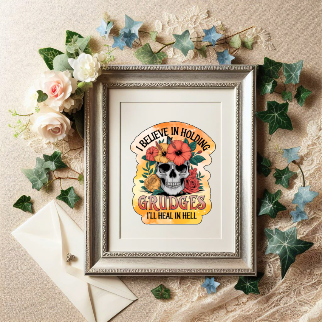 Holographic sticker with a floral-decorated skull and bright orange accents. Text reads “I Believe in Holding Grudges – I’ll Heal in Hell.” Inside a picture frame