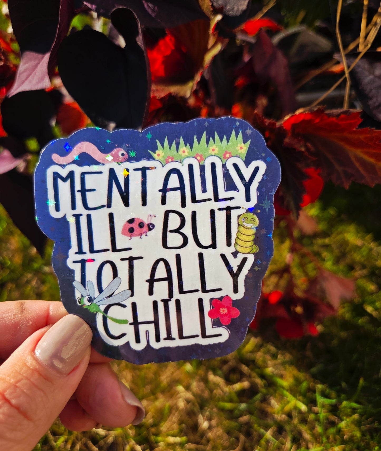 Holographic sticker that says “Mentally Ill But Totally Chill” with bugs, grass, and sparkles