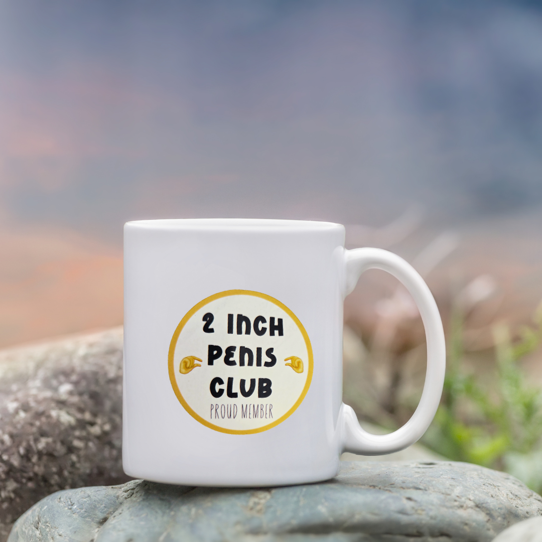 White mug with '2 Inch Penis Club' text on a rock with a blurred natural background