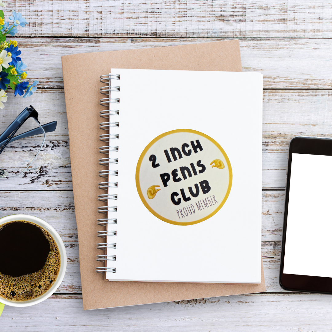 Notebook with '2 Inch Penis Club' sticker on a wooden surface with coffee and phone.