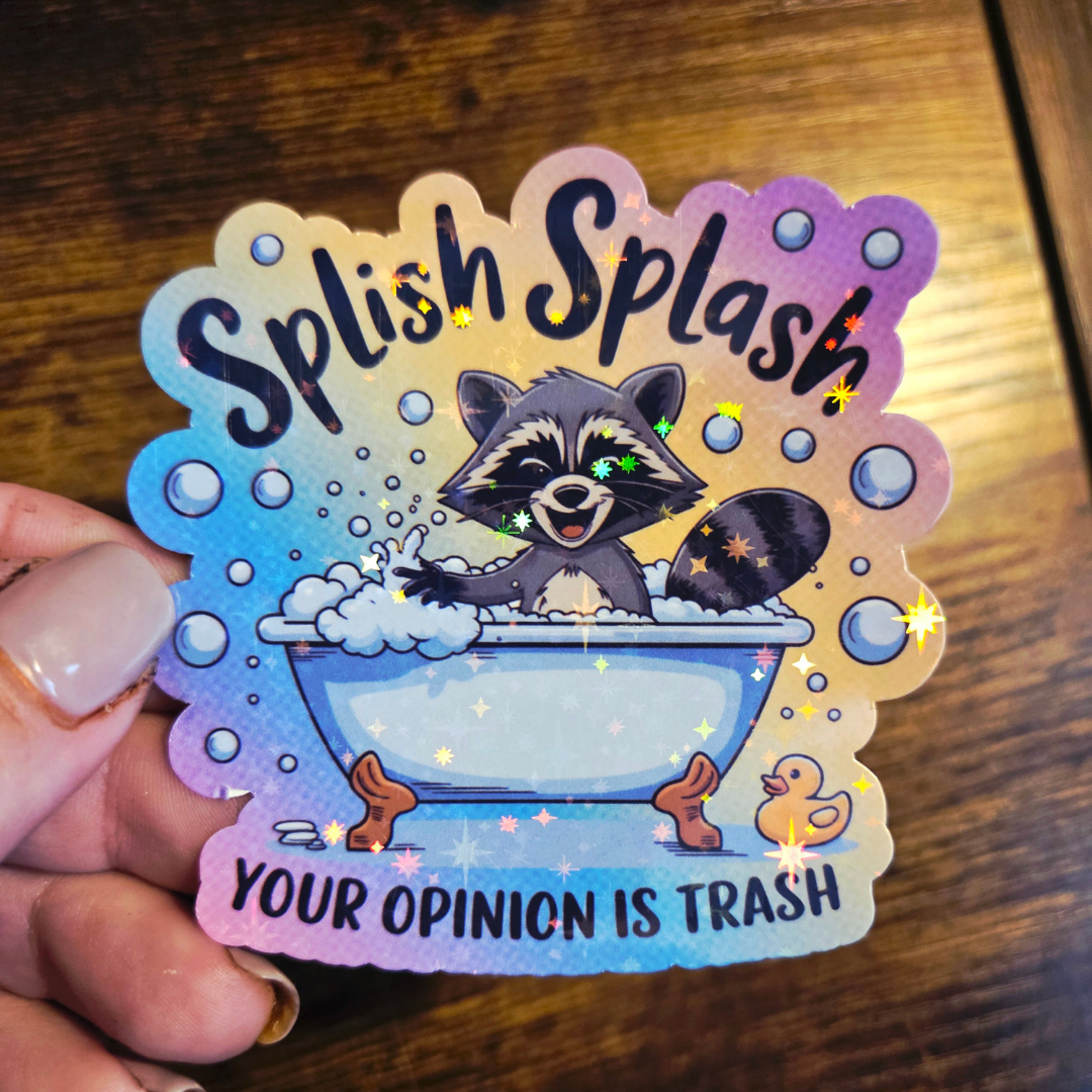 Sticker with a raccoon in a bathtub and 'Splish Splash Your Opinion is Trash' text on a wooden surface.