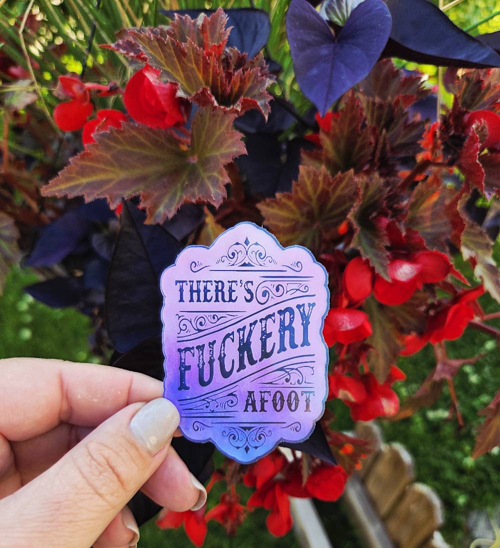 Sticker with ornate scrollwork and the phrase “There’s Fuckery Afoot” in black gothic-style lettering over a pink and purple gradient background