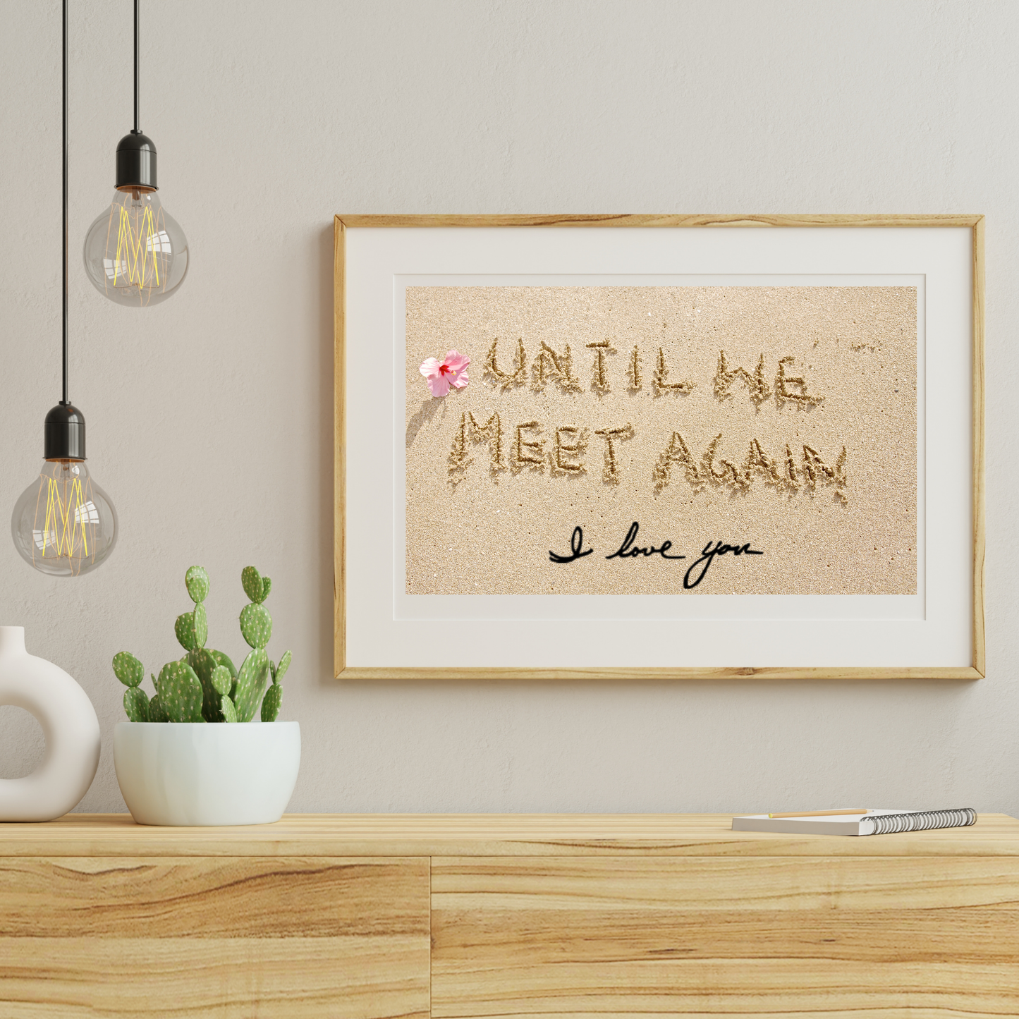Beach-themed framed print with the words “Until we meet again” written in sand, a small red cardinal, pink flower, and the handwritten message “I love you,” displayed above a light wooden dresser with hanging bulbs and cactus.