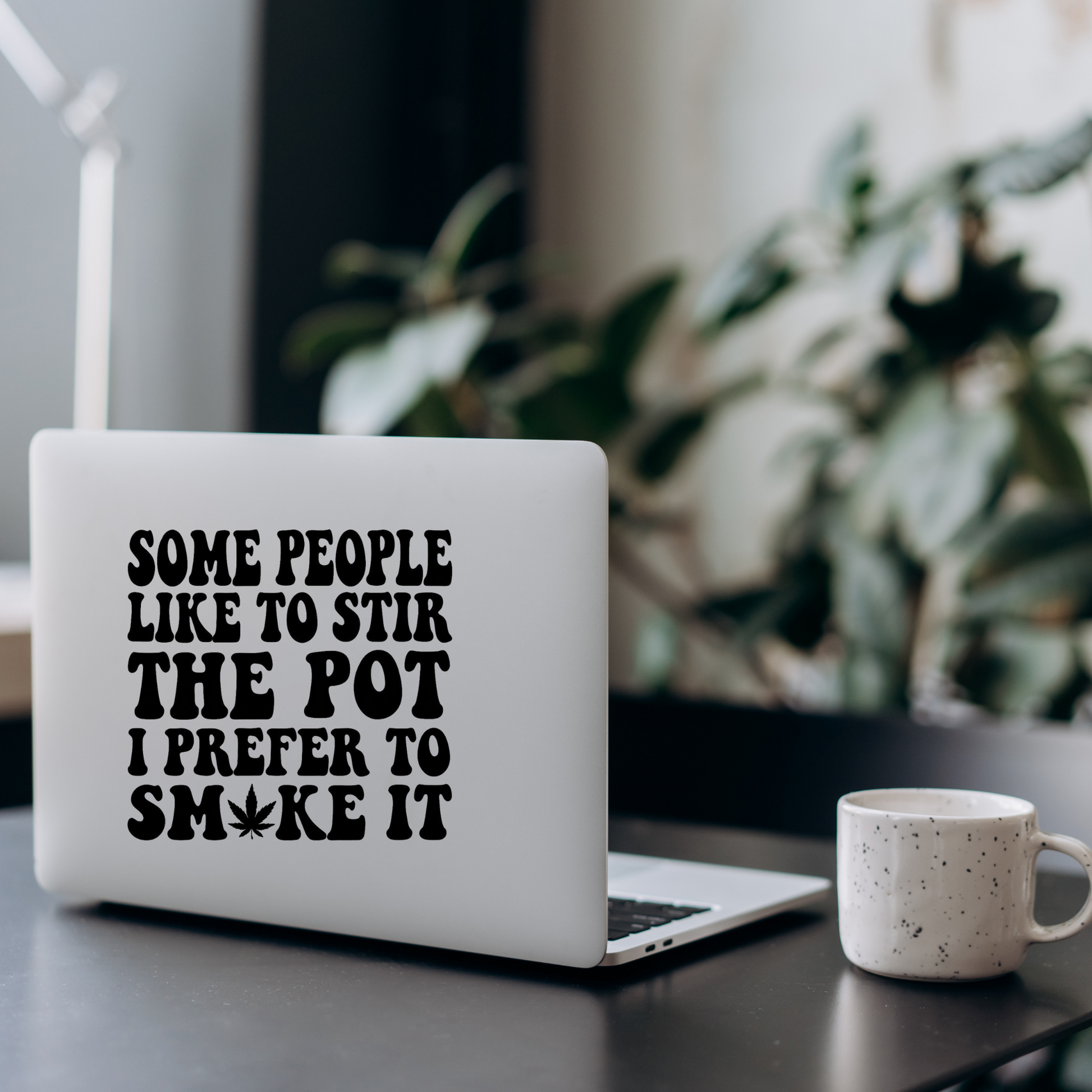 A laptop on a desk with a decal that says some people like to stir the pot i prefer to smoke it. Next to a coffee cup
