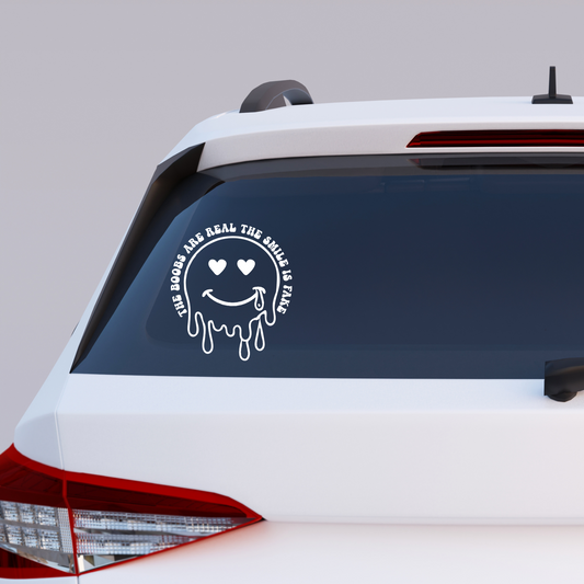 Vinyl decal of a dripping smiley face with heart eyes and a tear, surrounded by text that reads “The Boobs Are Real The Smile Is Fake”