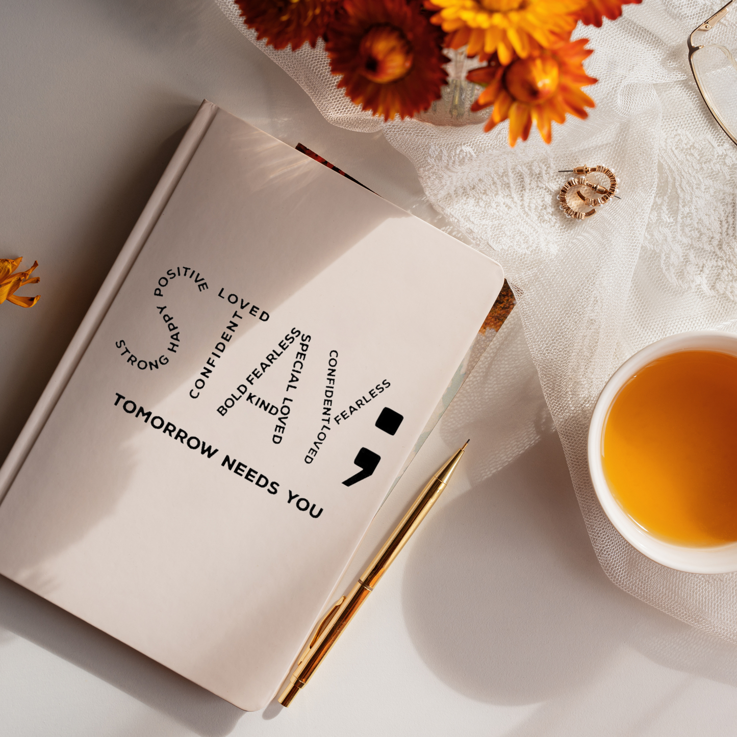 Beige journal with a decal that says stay; tomorrow needs you with the word "STAY" made up of other words like loved confident fearless. Orange flowers and a cup of tea in the background