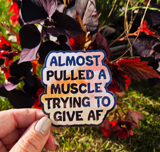 Holographic sticker that reads “Almost pulled a muscle trying to give AF” in bold black text over a peach-pink sparkle background