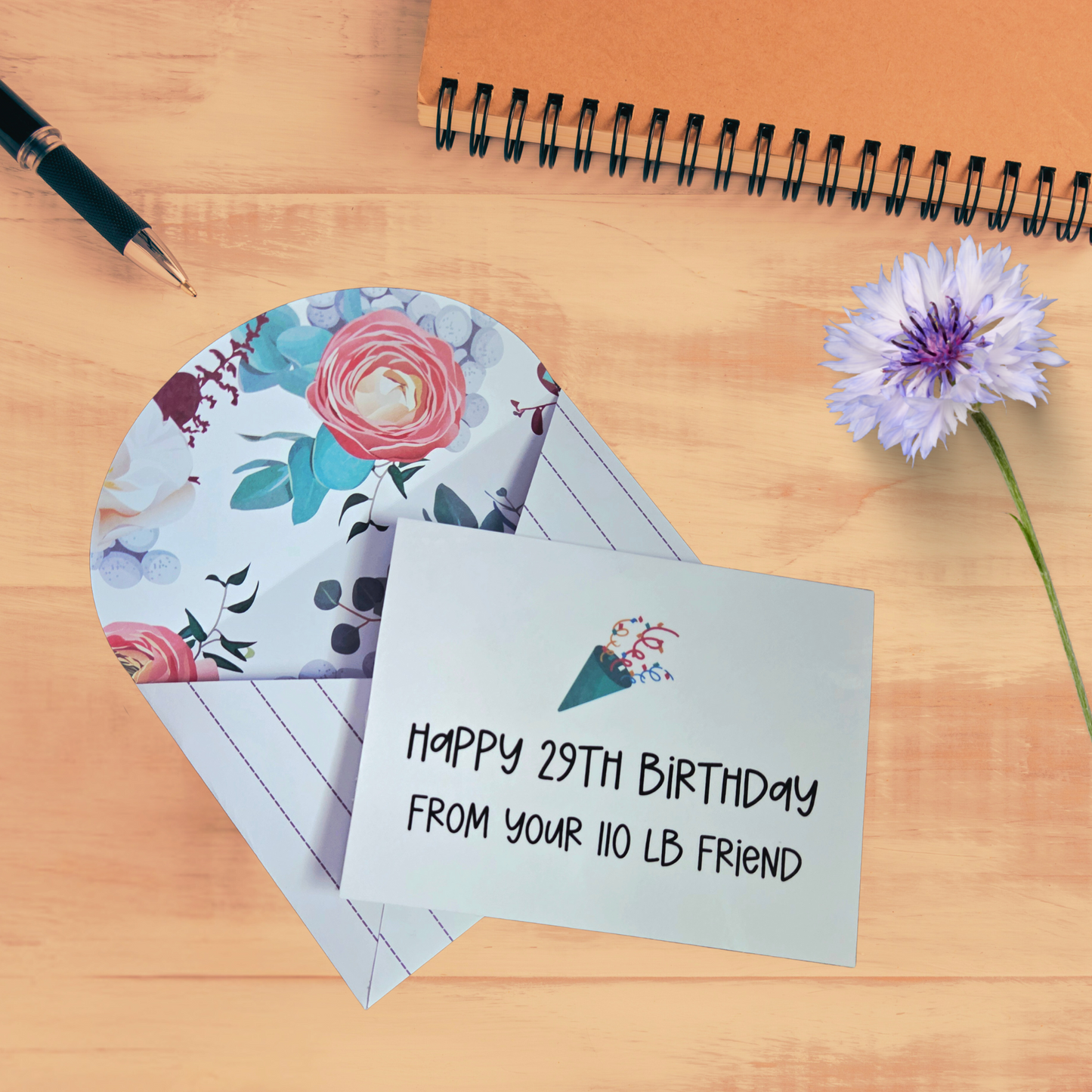 Birthday card with floral design and humorous message on a wooden surface with a pen and flower.
