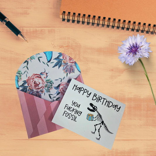 Funny 4x5.5 birthday card with dinosaur skeleton holding a gift that reads “Happy Birthday you f*cking fossil,” shown with handmade floral envelope.