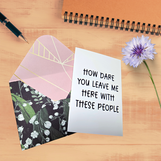 Greeting card with text 'How dare you leave me here with these people' on a wooden surface with an envelope and flower.