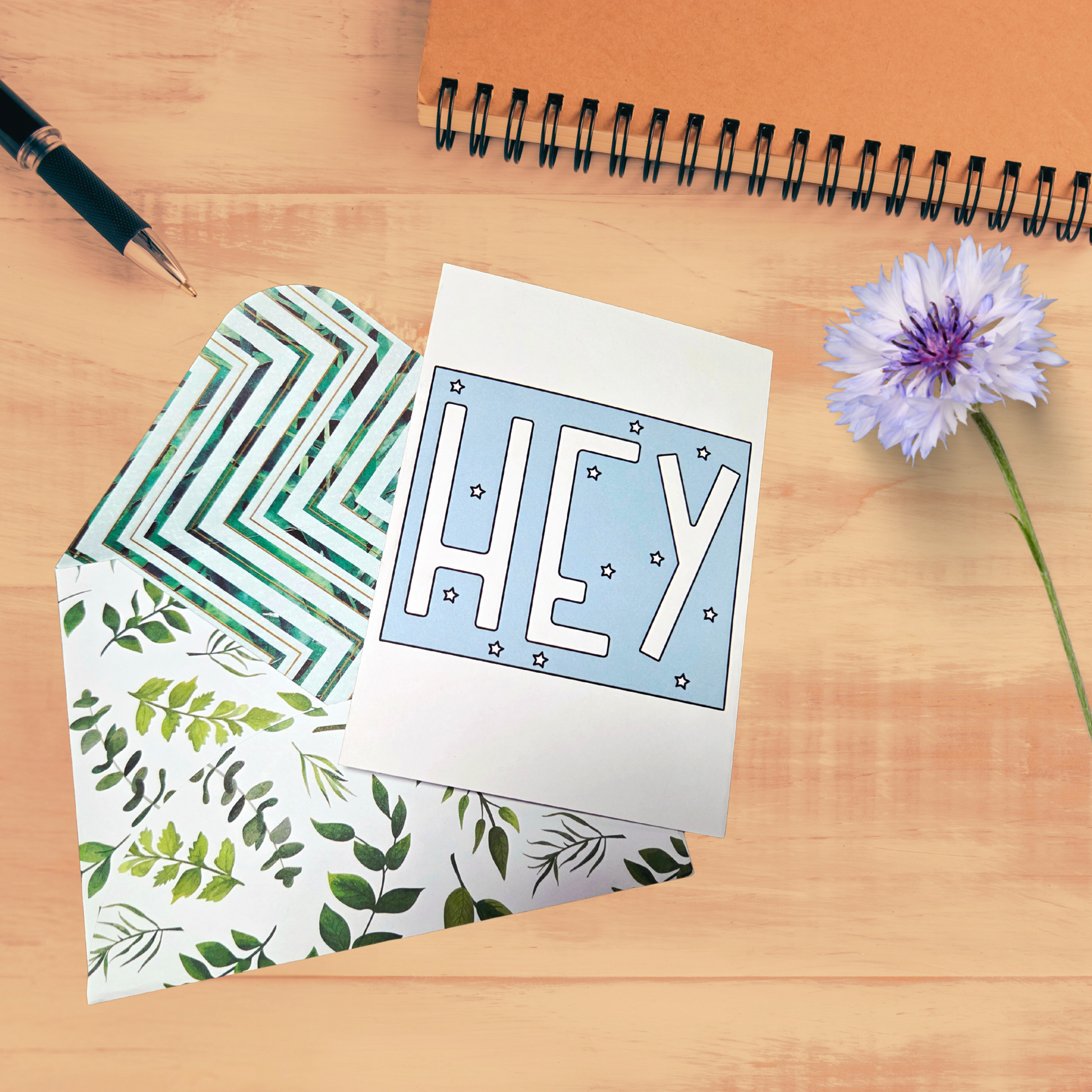 Greeting card with 'HEY' text, patterned envelope, pen, and flower on a wooden surface