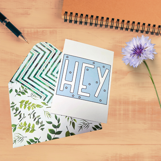 Greeting card with 'HEY' text, patterned envelope, pen, and flower on a wooden surface