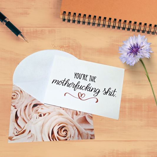Card with text 'You're the motherfucking shit' on a wooden surface with a flower and pen.