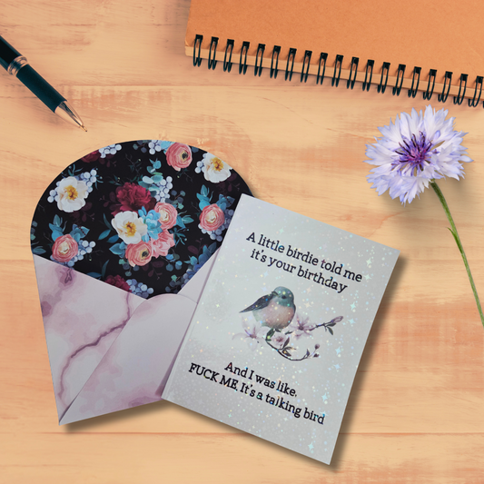 Greeting card with floral design and humorous text on a wooden surface with a pen and flower.