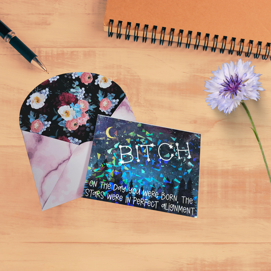 Greeting card with holographic starry design reading “BITCH – On the day you were born, the stars were in perfect alignment,” paired with a floral-lined envelope. 