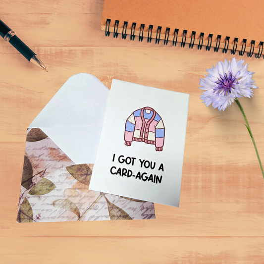 Greeting card with a colorful cardigan illustration and text on a wooden surface with a pen and flower.