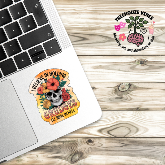 Holographic sticker with a floral-decorated skull and bright orange accents. Text reads “I Believe in Holding Grudges – I’ll Heal in Hell.”