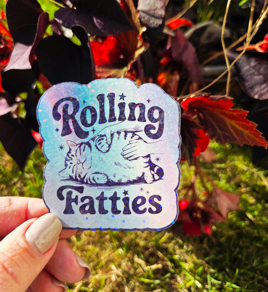 Holographic sticker with a hand rubbing a fat, relaxed cat and the phrase “Rolling Fatties” in playful black text