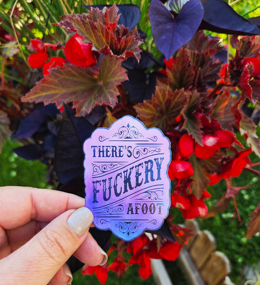 Sticker with ornate scrollwork and the phrase “There’s Fuckery Afoot” in black gothic-style lettering over a pink and purple gradient background