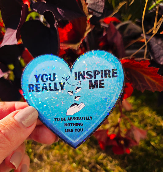 Heart-shaped holographic sticker with the phrase “You really inspire me to be absolutely nothing like you” on a blue sparkle background